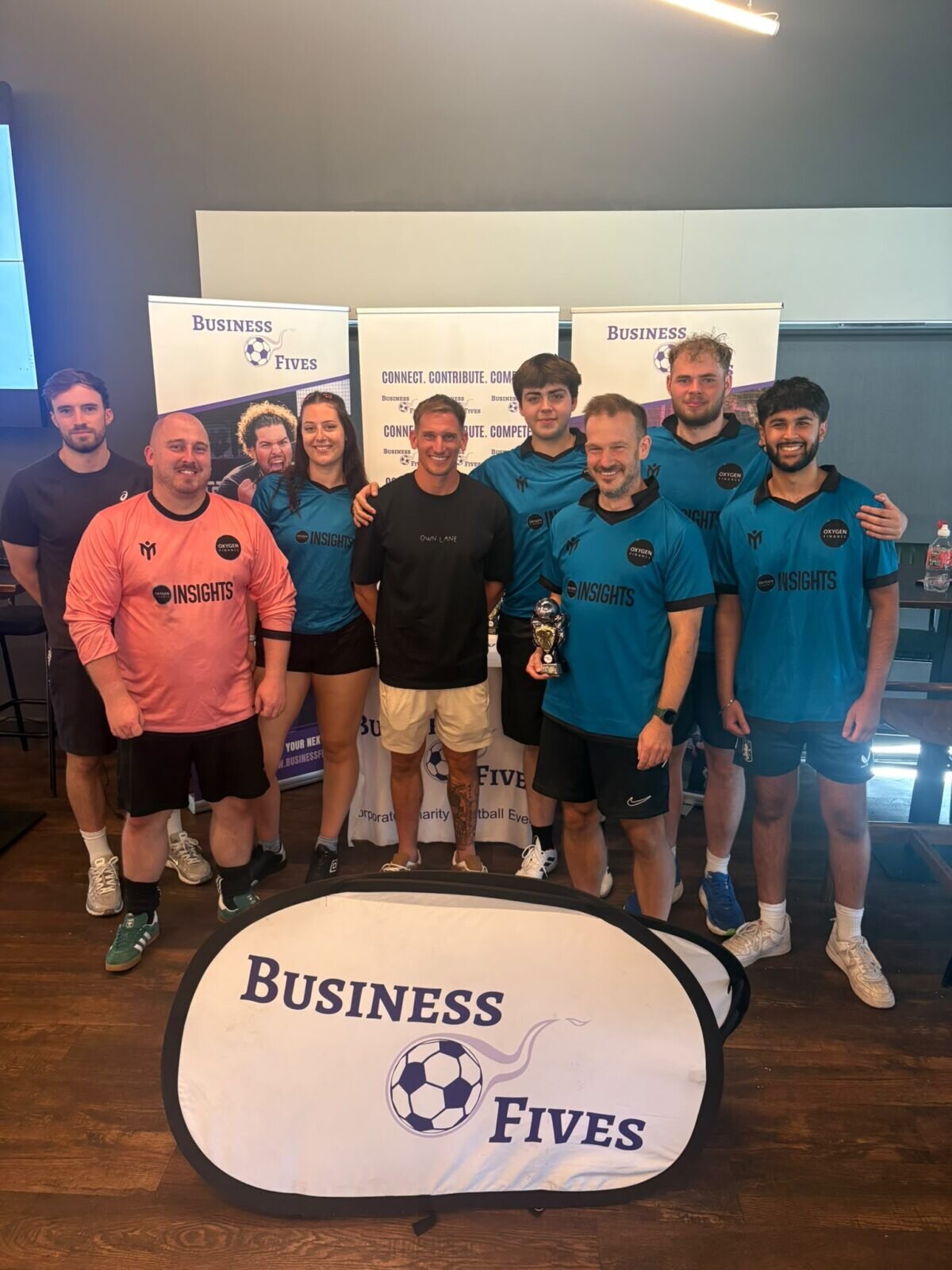 Nine of us in teal kit and one keeper in pink, holding a small trophy in front of Business Fives banners