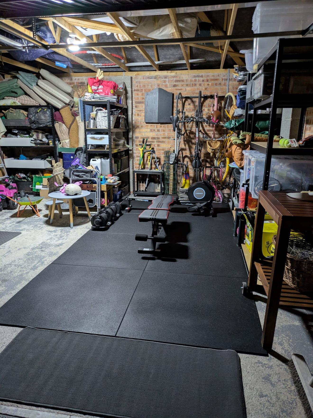 The cleared corner of the garage set up as a gym: rubber floor mats, a weights bench, adjustable dumbbells and an exercise bike