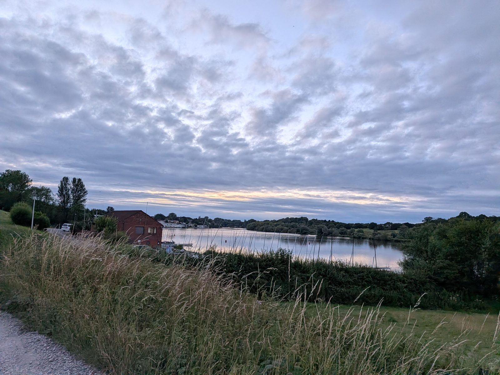 Winsford flash at dusk, still water and moored sailing boats under a grey and gold evening sky, tall grass in the foreground