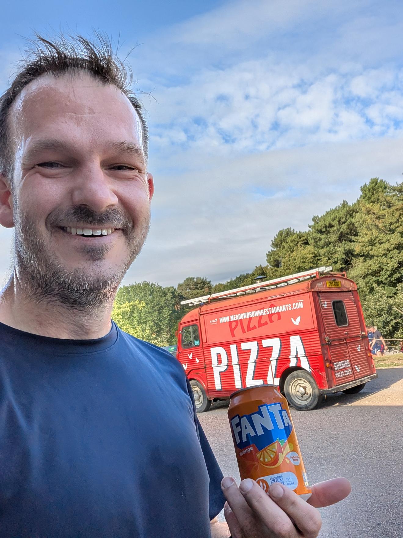 Selfie after the time trial, grinning, holding a can of Fanta, with a red pizza van parked in the background