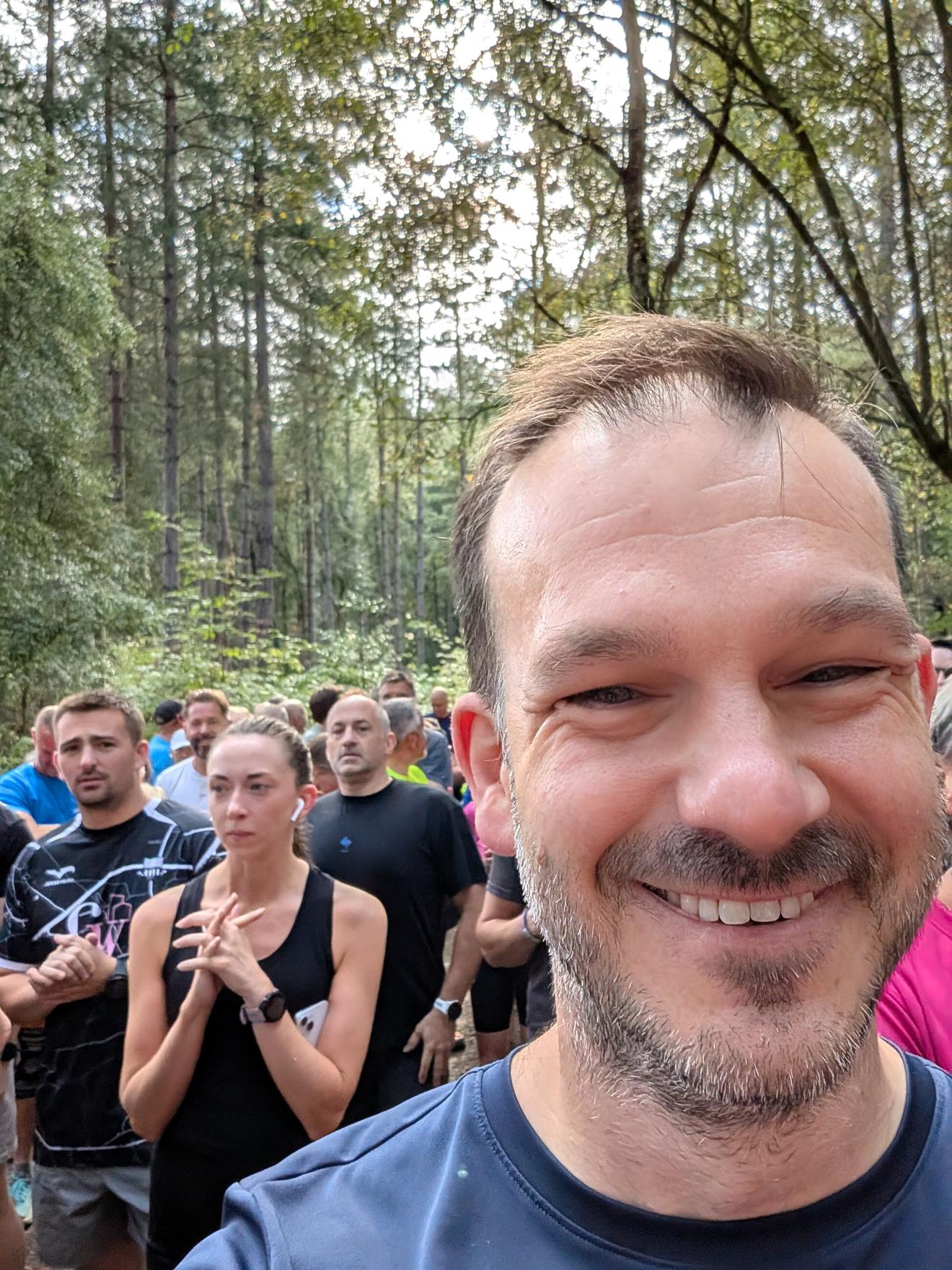 Selfie at the Delamere parkrun start line, grinning, with a crowd of runners waiting behind among the pine trees