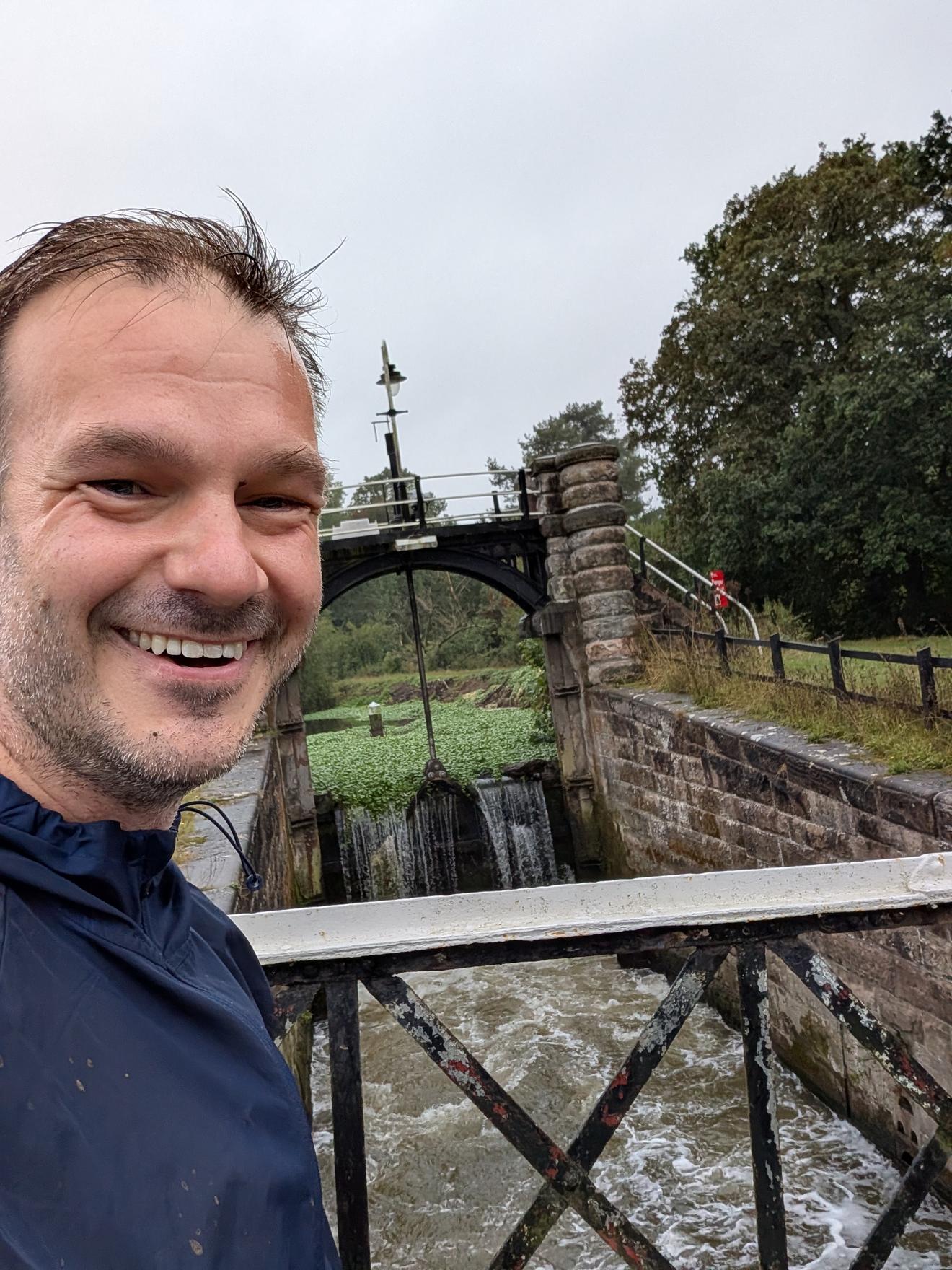 Selfie on the old iron footbridge over a lock on the River Weaver, rain soaked, water rushing over the weir behind