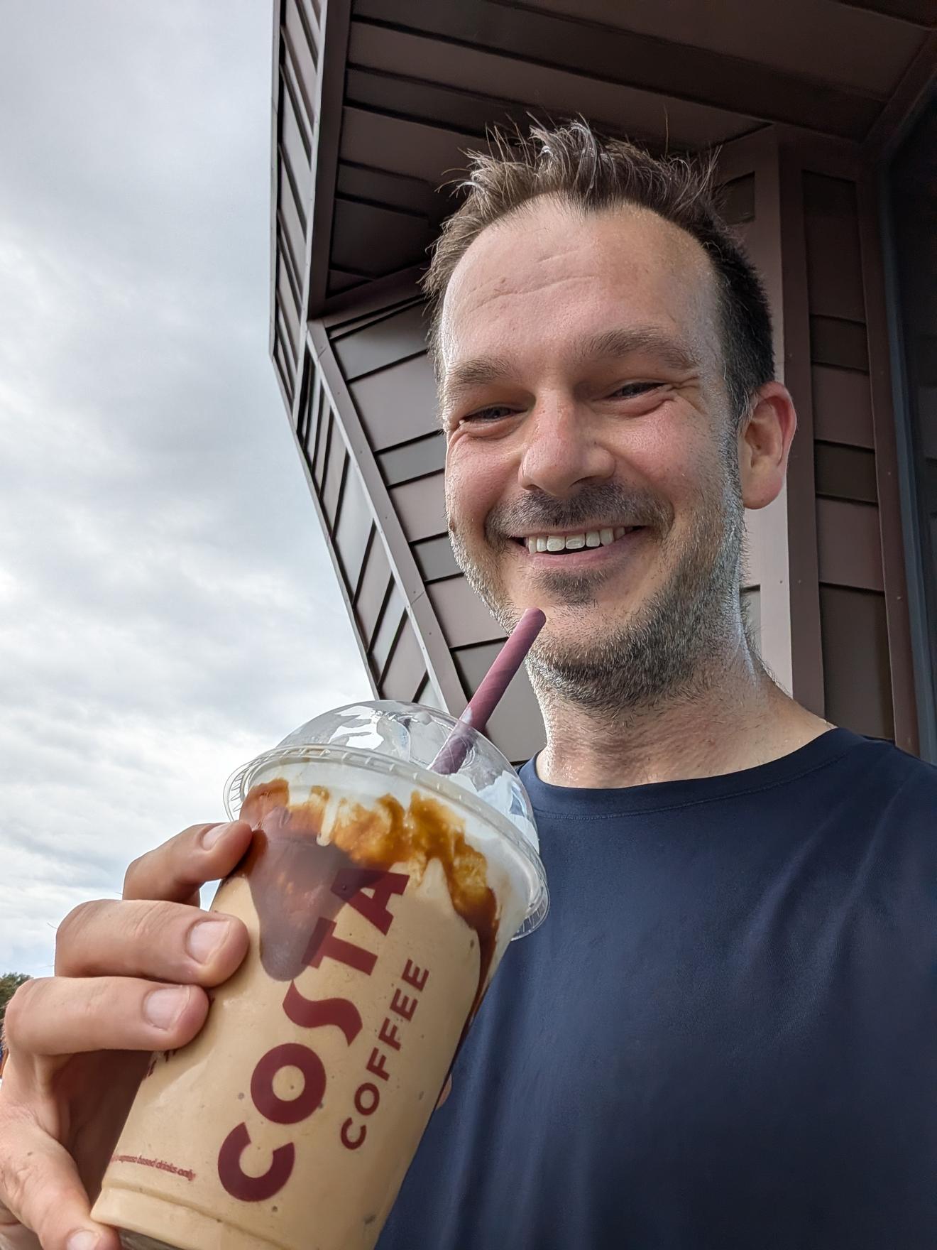 Holding an iced Costa Frappe with caramel drizzle, grinning, still in running kit